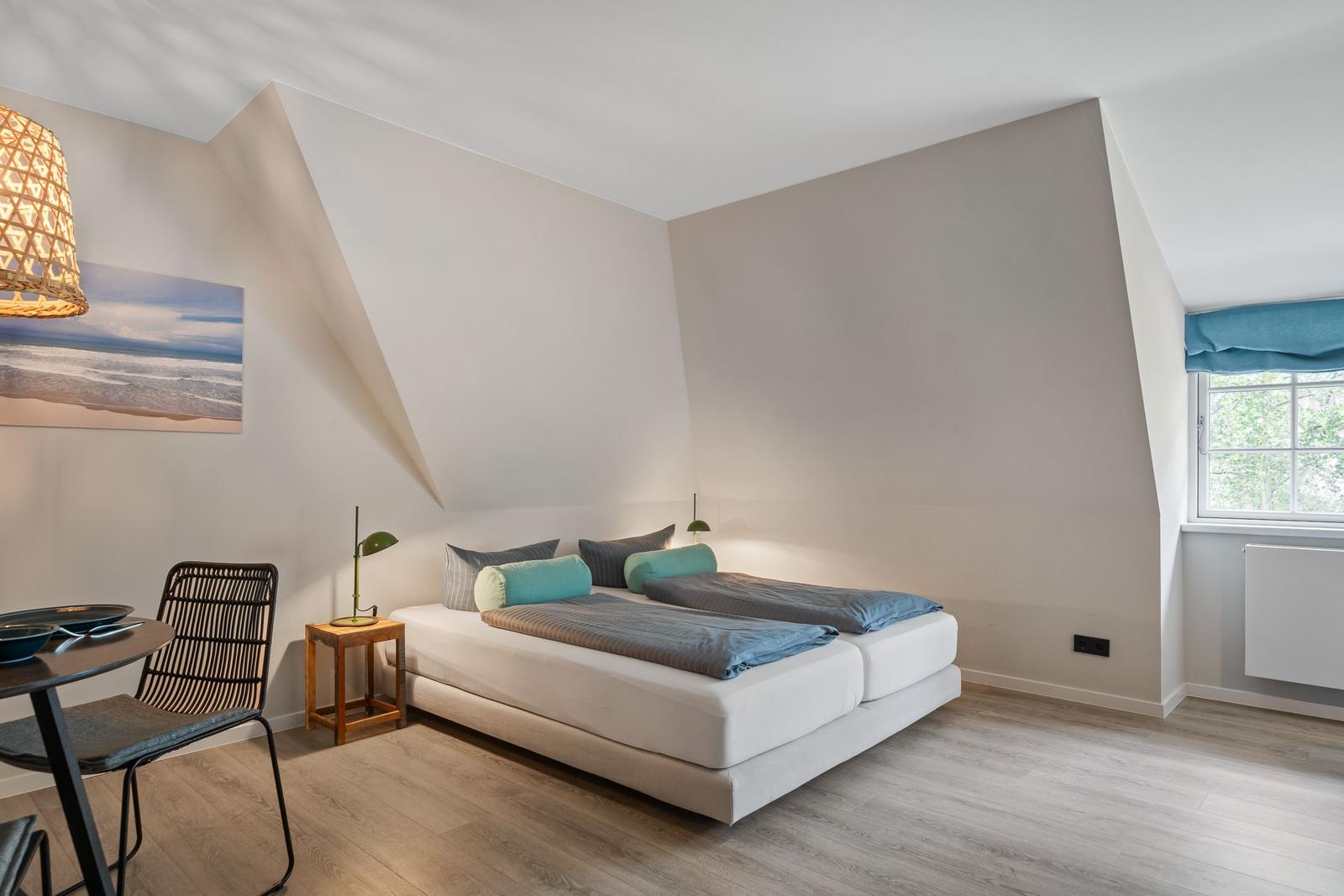 Bedroom with double bed, window, and dining area. Wooden floor and wall art.
