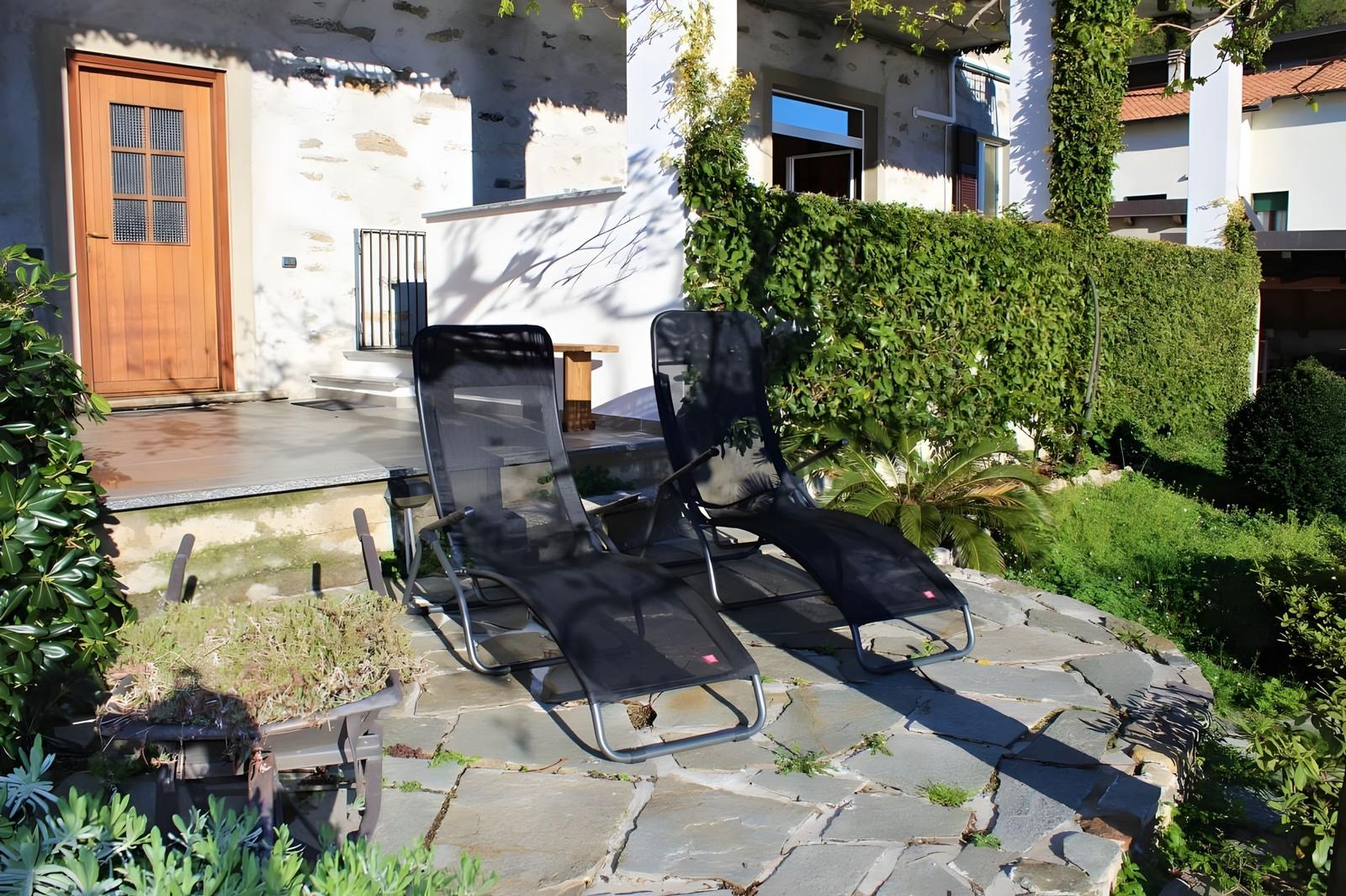 Two black lounge chairs on stone pavers in front of a house with green hedge.
