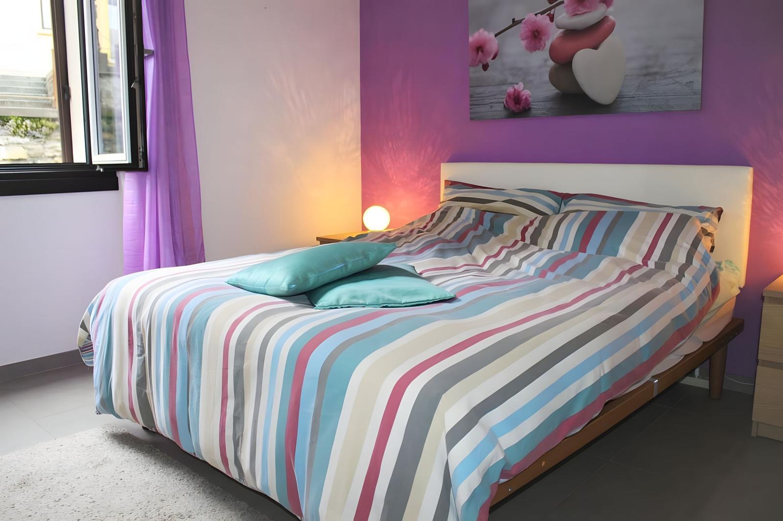 Bedroom with bed, wall art, and window. Purple walls, bed with striped bedding.