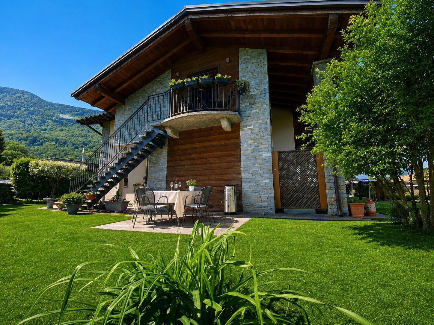 Haus mit Balkon, Terrasse und Garten. Hintergrund: bewaldeter Berg.