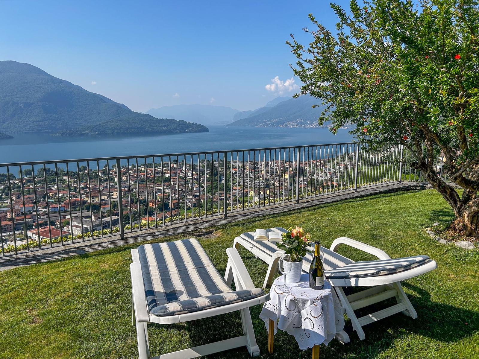 Terrasse mit Liegestühlen und Tisch. Blick auf See und Stadt. Grasfläche und Baum.