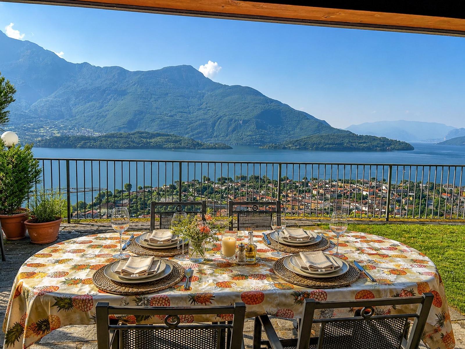Runder Tisch mit Tischdecke und Stühlen auf Terrasse mit Blick auf See und Berge.