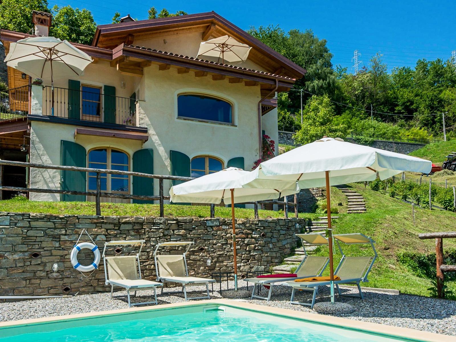 Poolbereich mit Liegestühlen und Sonnenschirmen vor einer Villa mit Steinmauer.