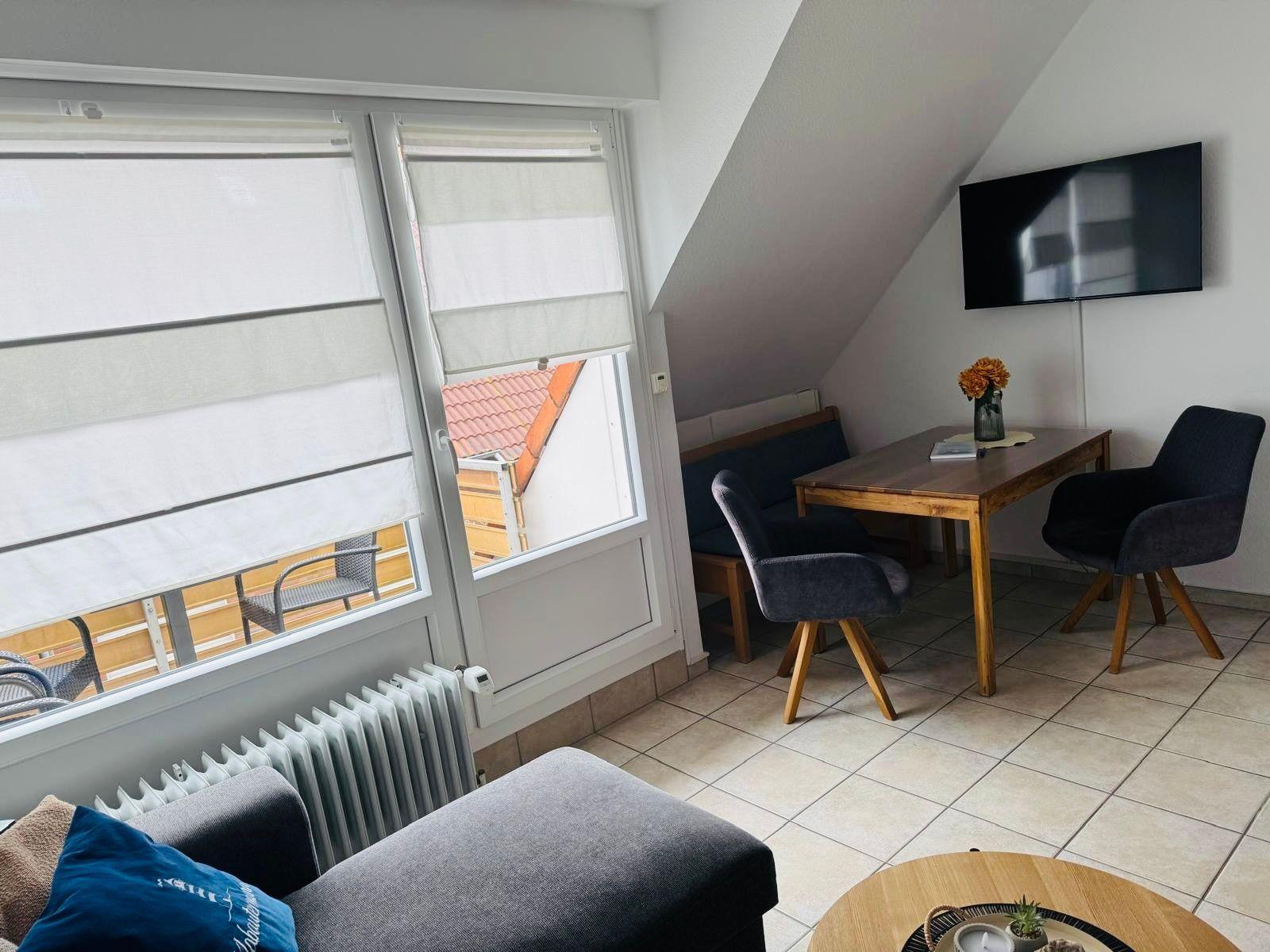 Living area with dining table, TV, and balcony view. Wooden table, chairs, and sofa.