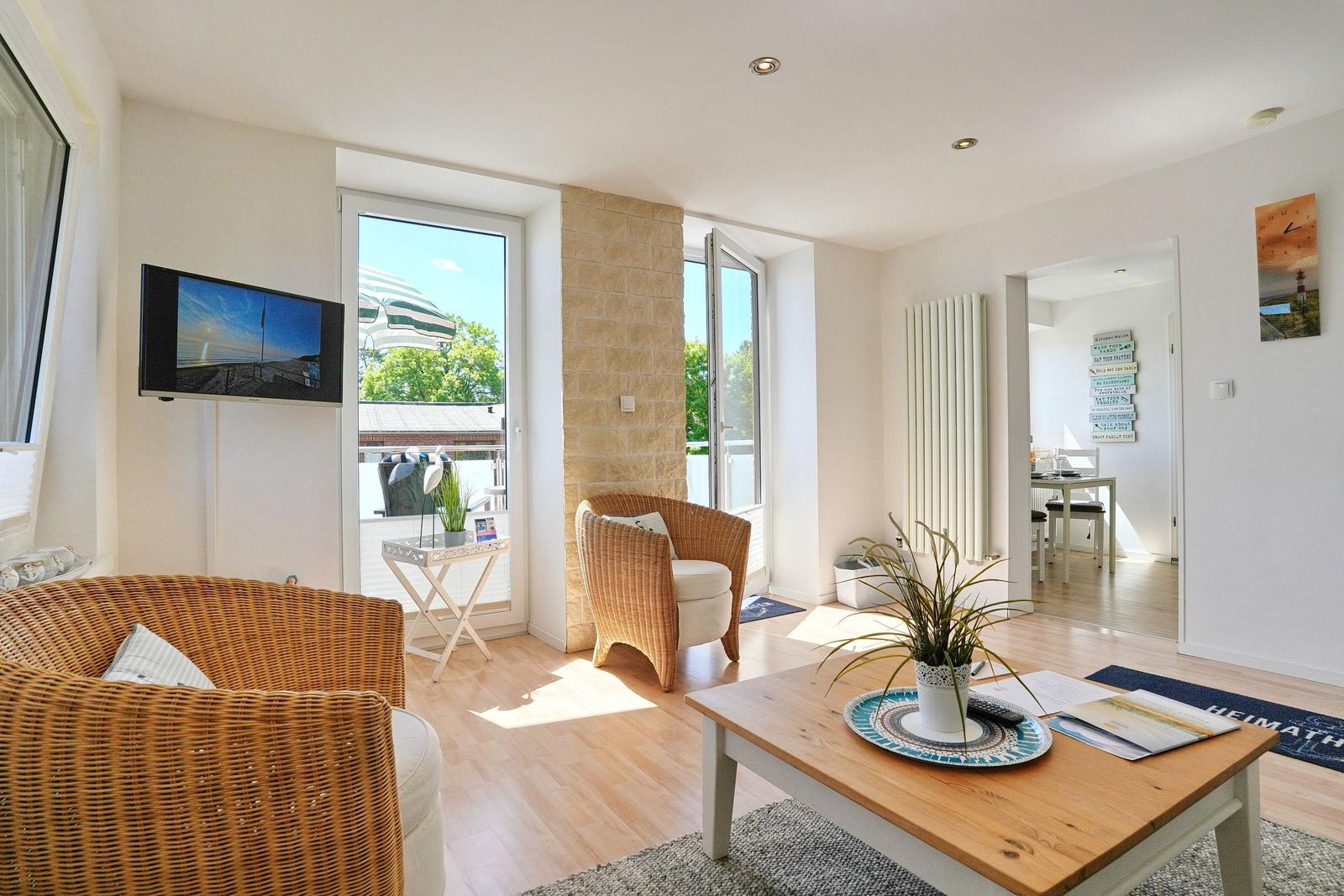 Wohnzimmer mit Sitzgelegenheiten, TV und Blick auf Balkon. Essbereich im Hintergrund.