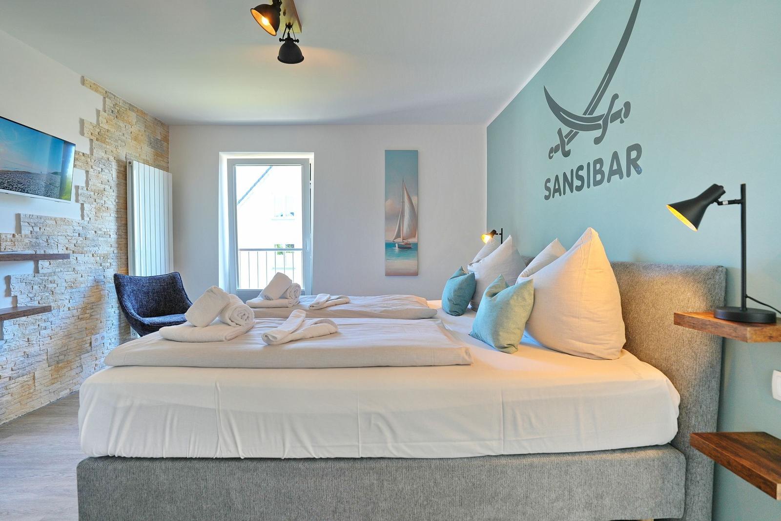 Zimmer mit Doppelbett, Wanddekoration und Blick auf Balkon.