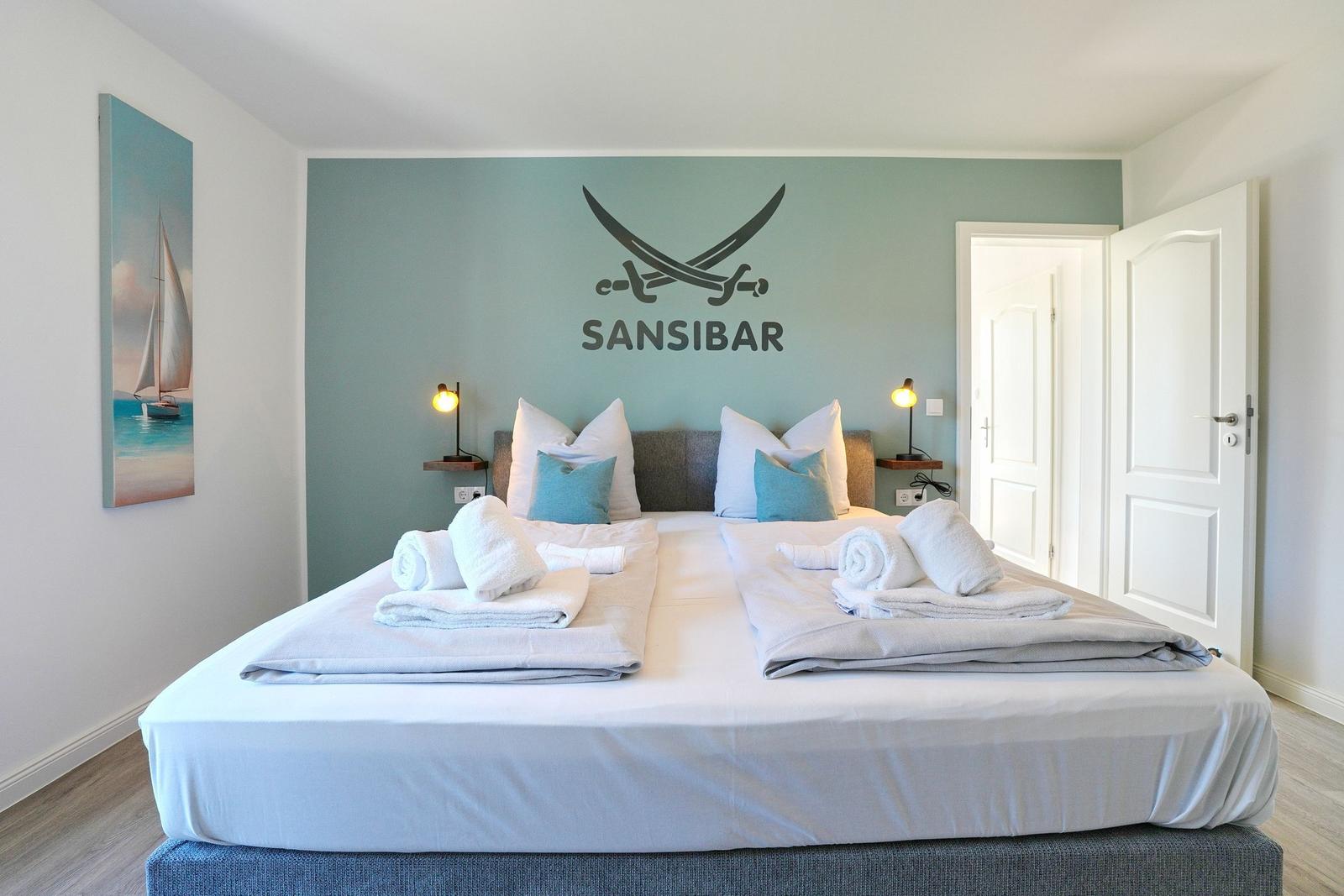 Doppelbett mit weißen Laken, blauen Kissen und gefalteten Handtüchern. Wand mit Logo und Segelboot-Bild.