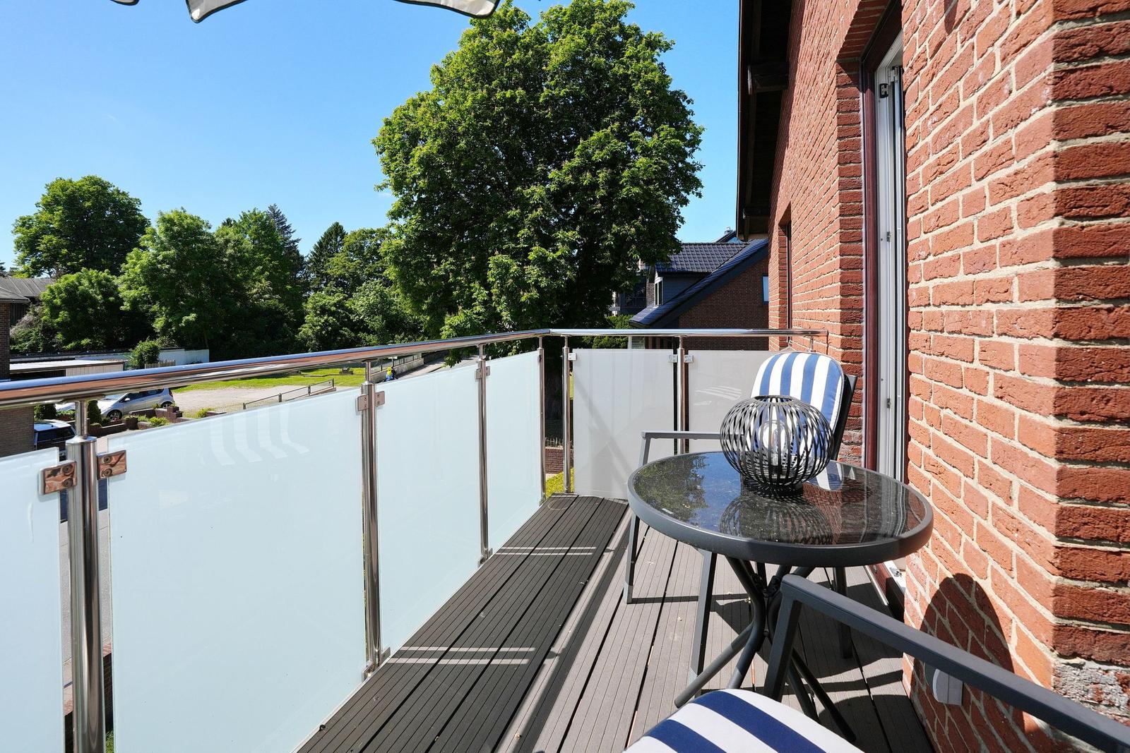 Terrasse mit Tisch, Stühlen und Glasgeländer. Blick auf Bäume und Gebäude.