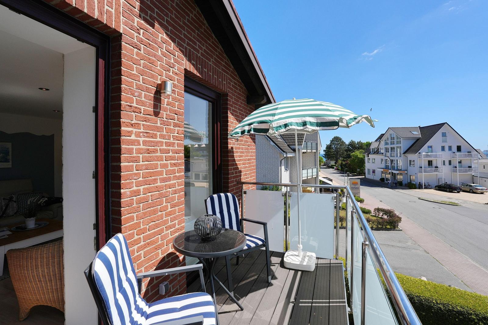 Terrasse mit Tisch, zwei Stühlen und Sonnenschirm. Blick auf Straße und Gebäude.