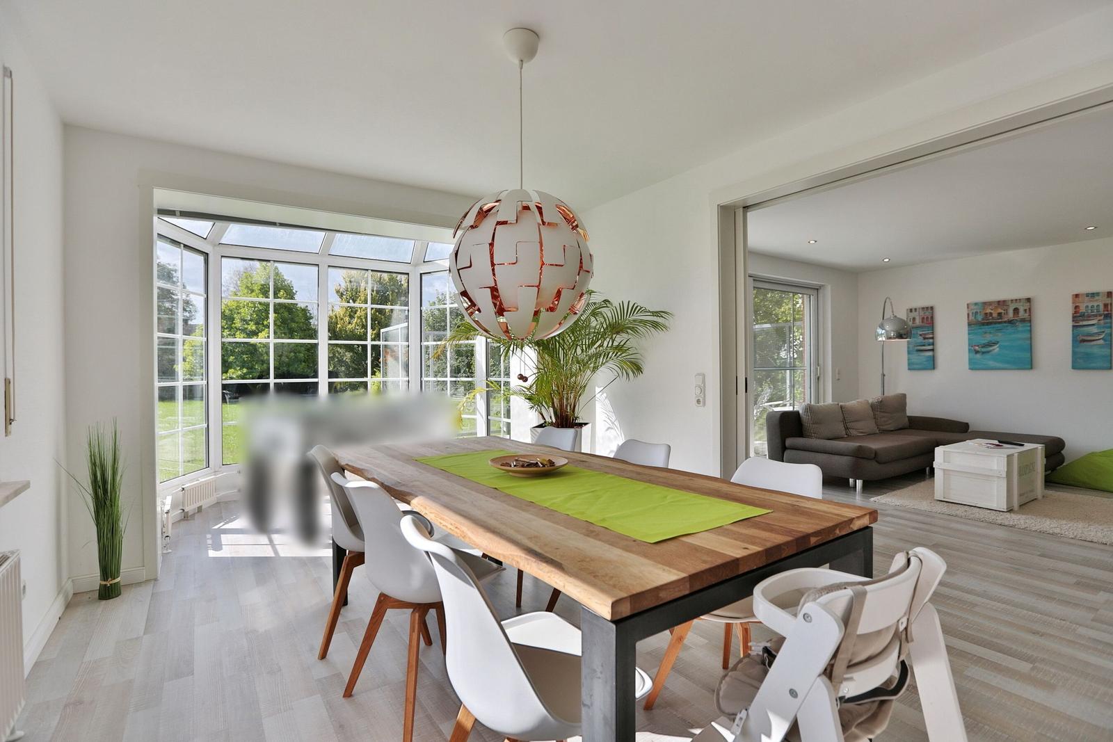 Essbereich mit Holztisch, weißen Stühlen und großem Fenster. Wohnzimmer mit Sofa und Wandbildern.