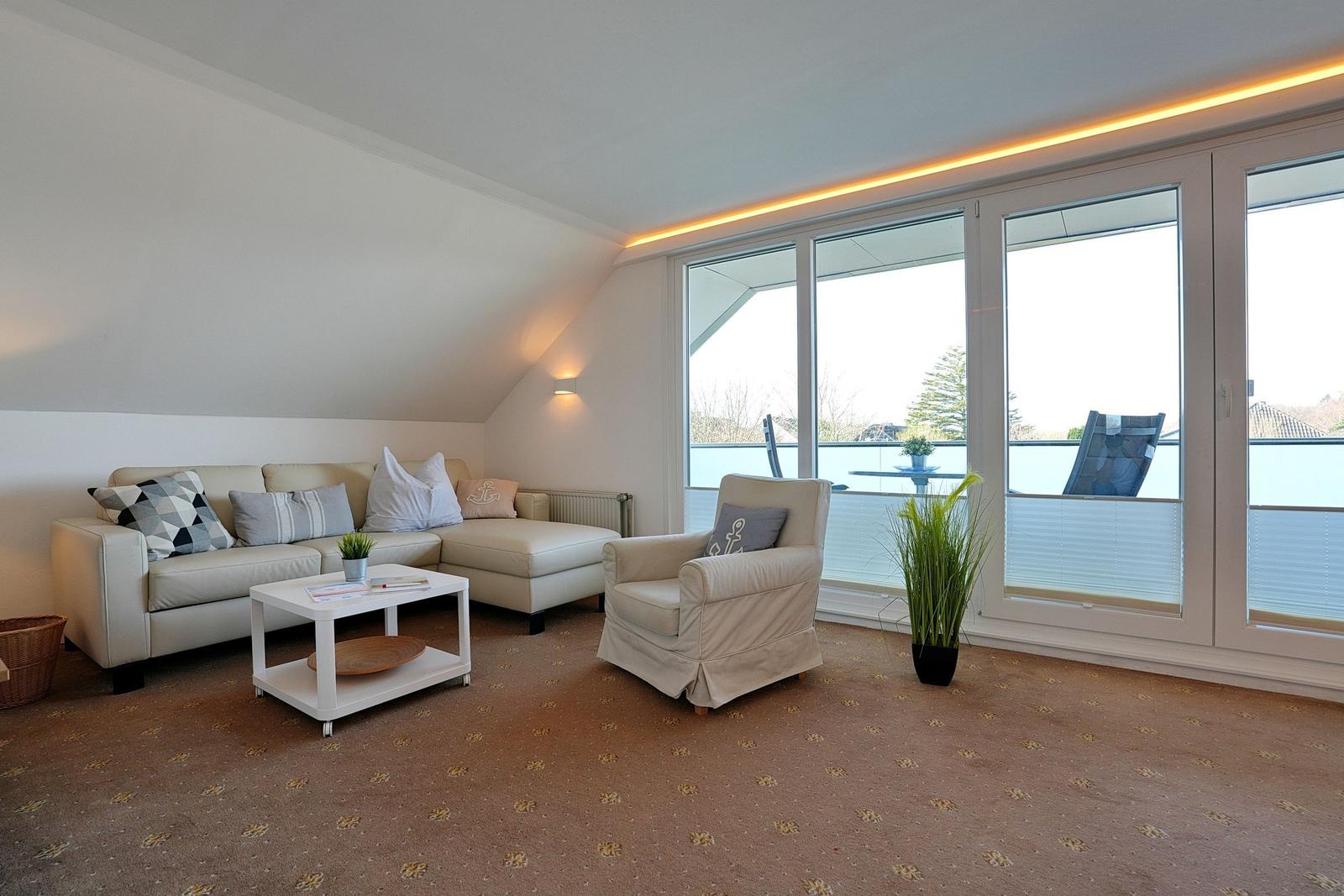 Wohnzimmer mit Sofa, Sessel und Balkonansicht. Teppichboden und große Fenster.
