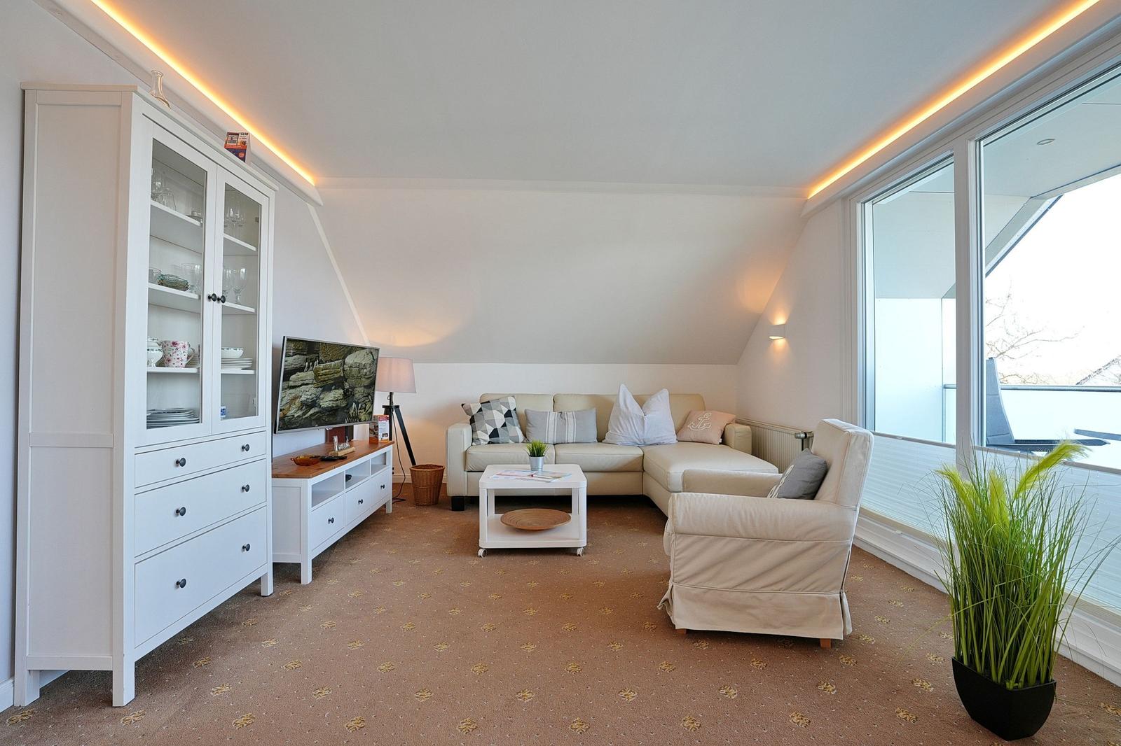 Wohnzimmer mit Sofa, Sessel, Fernseher und Glasvitrine. Große Fenster und Teppichboden.