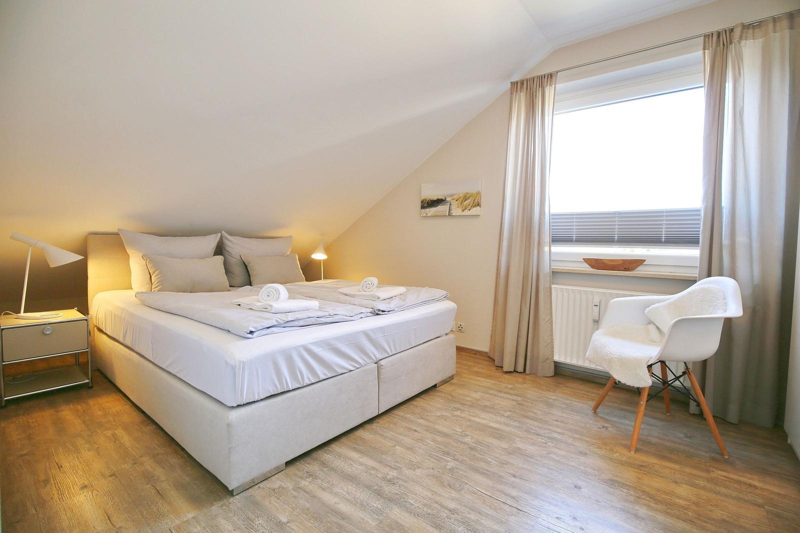 Schlafzimmer mit Doppelbett, Nachttisch, Stuhl und Fenster mit Vorhängen.