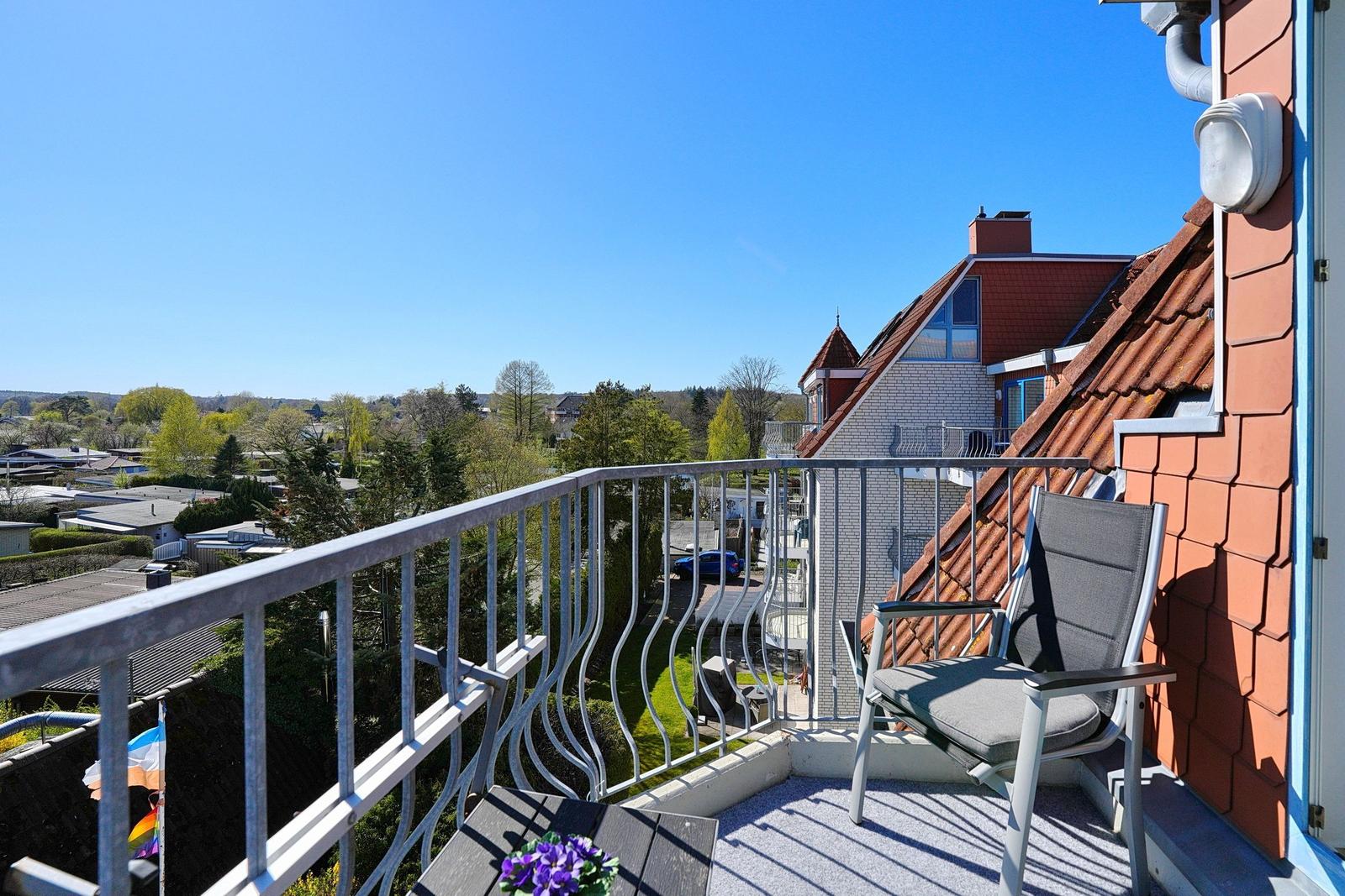Balkon mit Stuhl und Tisch, Blick auf Dächer und Bäume unter blauem Himmel.