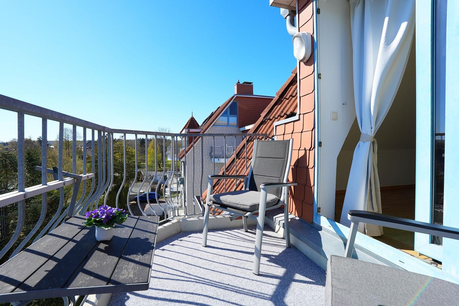 Balkon mit Stuhl, Tisch und Blumen. Blick auf Dach und Grün.