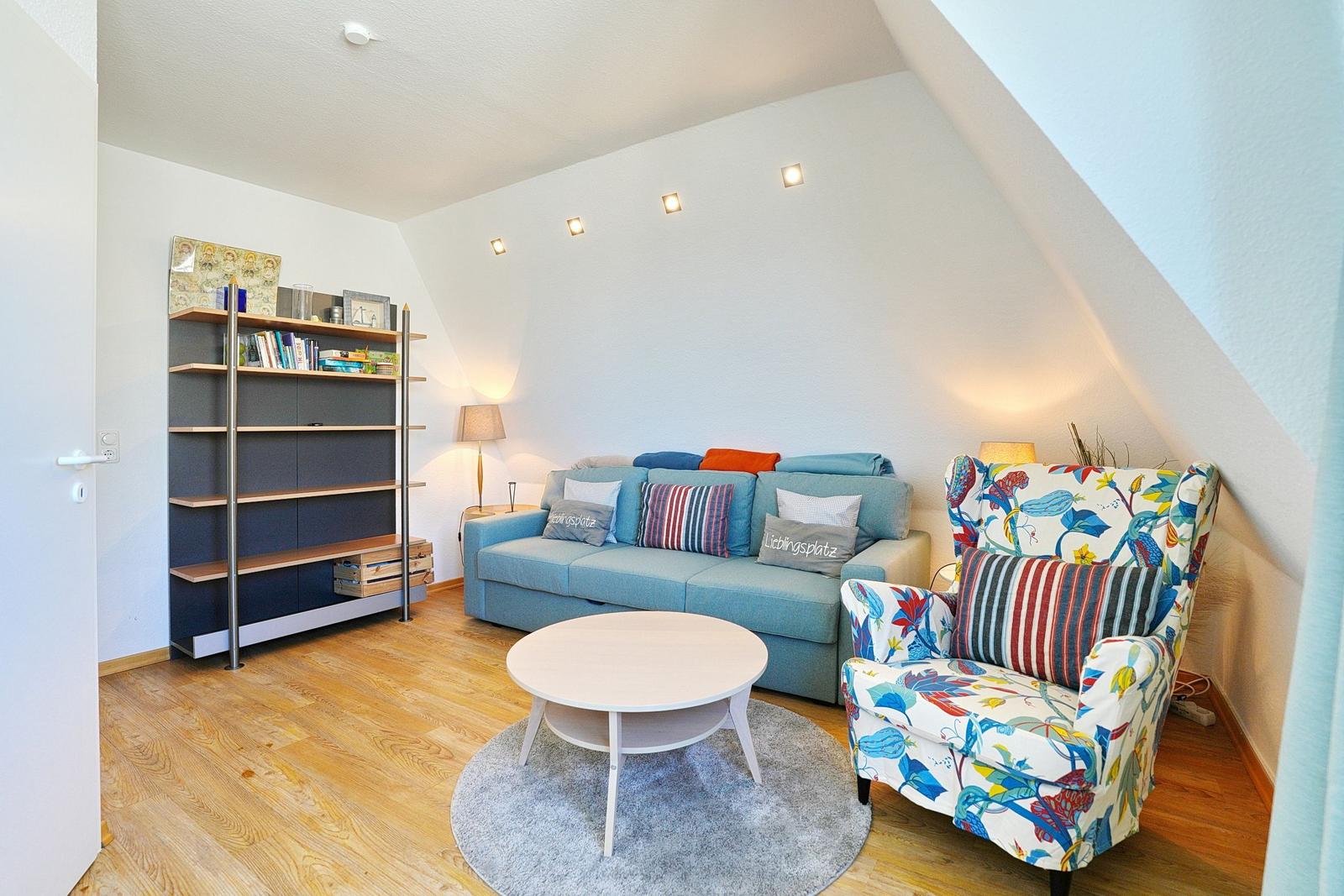 Wohnzimmer mit Sofa, Sessel und Regal unter schrägen Dachdecken.