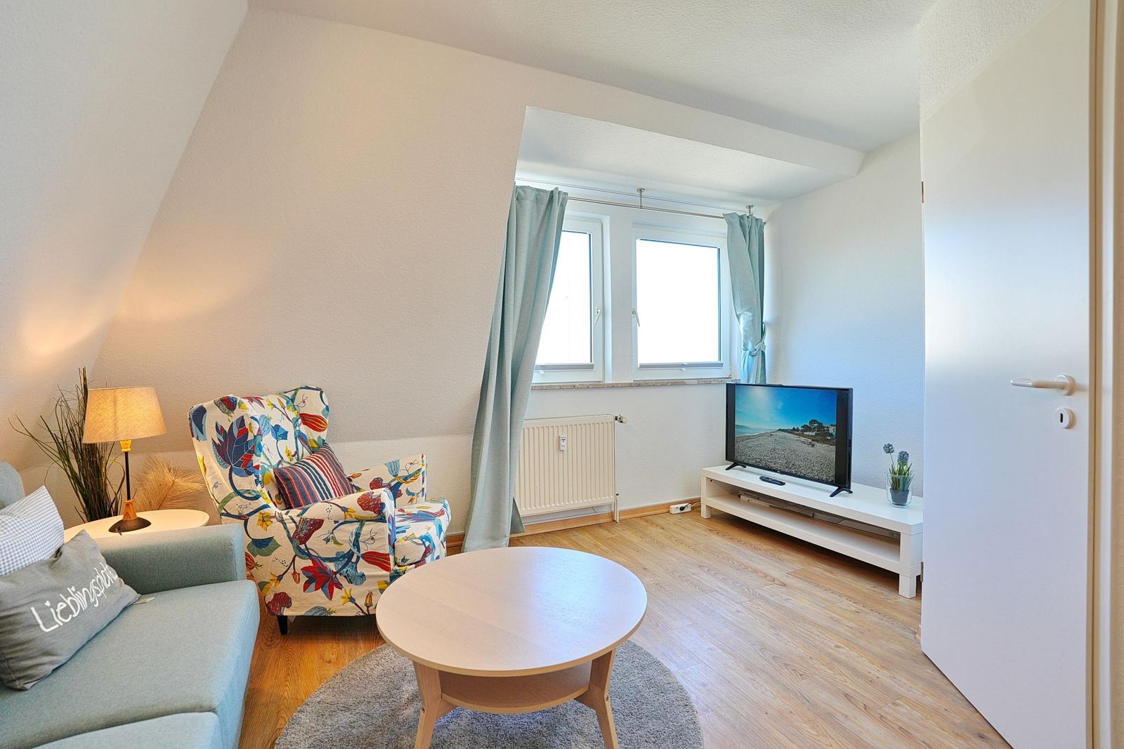 Wohnzimmer mit Sitzgelegenheiten, Fernseher und Fenster mit Vorhängen.