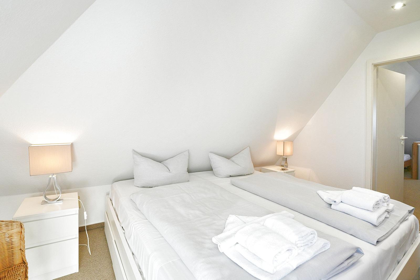 Schlafzimmer mit Doppelbett, zwei Nachttischen und Lampen. Weiße Wände und schräge Decke.