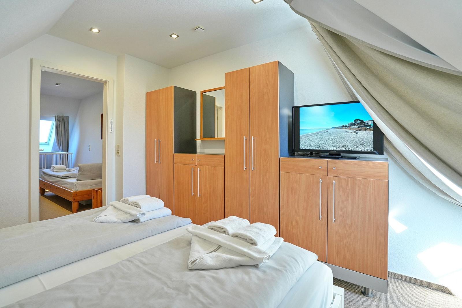 Zimmer mit zwei Betten, Schrank und Fernseher. Blick in angrenzendes Zimmer.