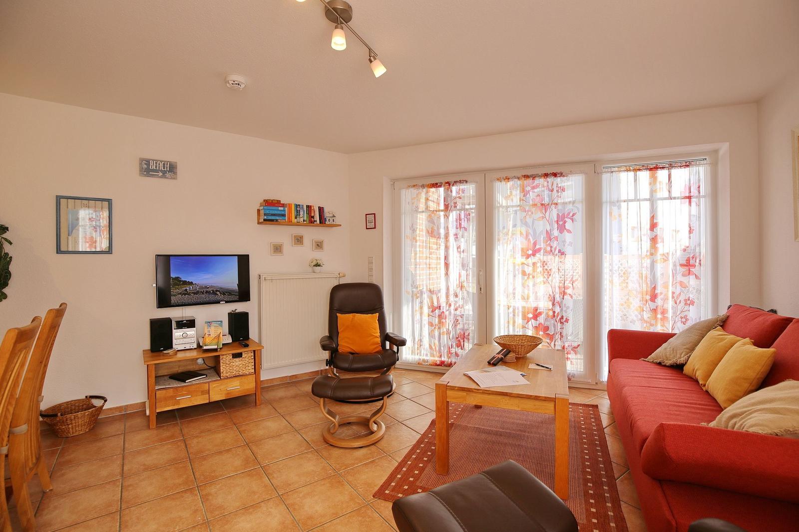 Wohnzimmer mit rotem Sofa, Holztisch und Fernseher. Glastüren führen auf eine Terrasse.