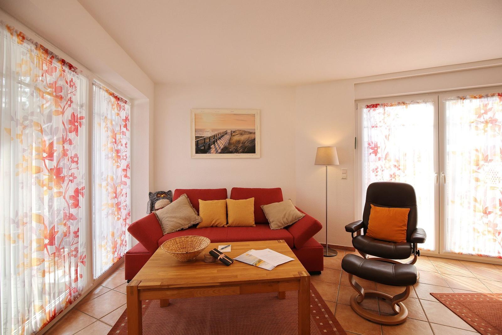 Ein Wohnzimmer mit rotem Sofa, Holztisch und Sessel. Fenster mit floralen Vorhängen.