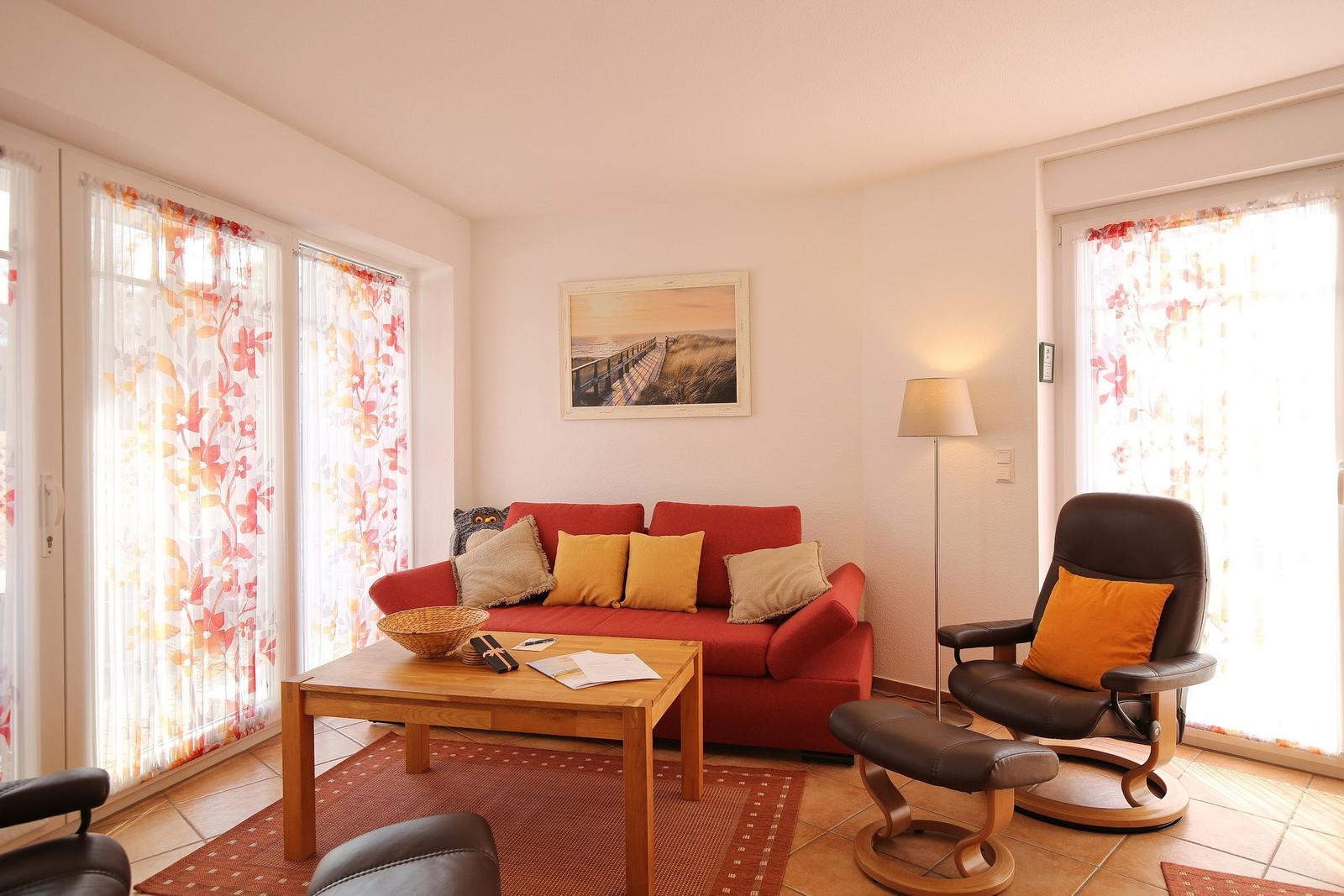 Ein Wohnzimmer mit rotem Sofa, braunem Sessel und Holztisch. Fenster mit floralen Vorhängen.