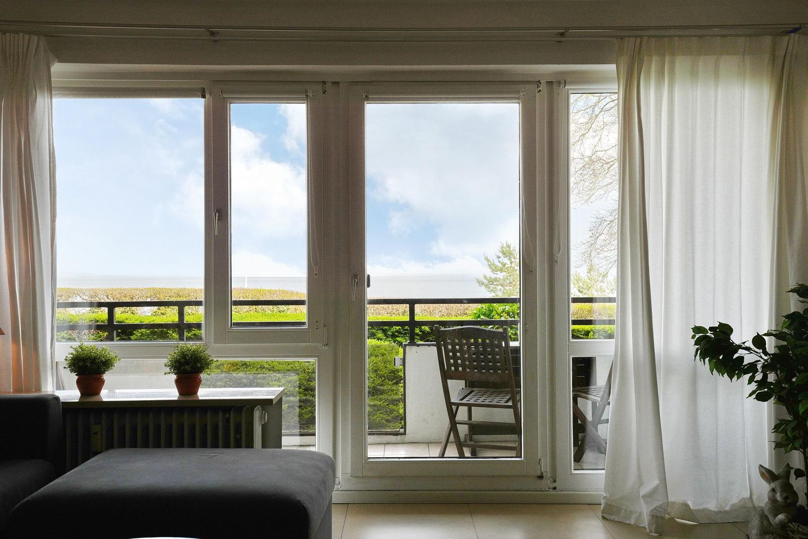 Durch das Fenster blickt man auf einen Balkon mit Stuhl und Blick auf das Meer.