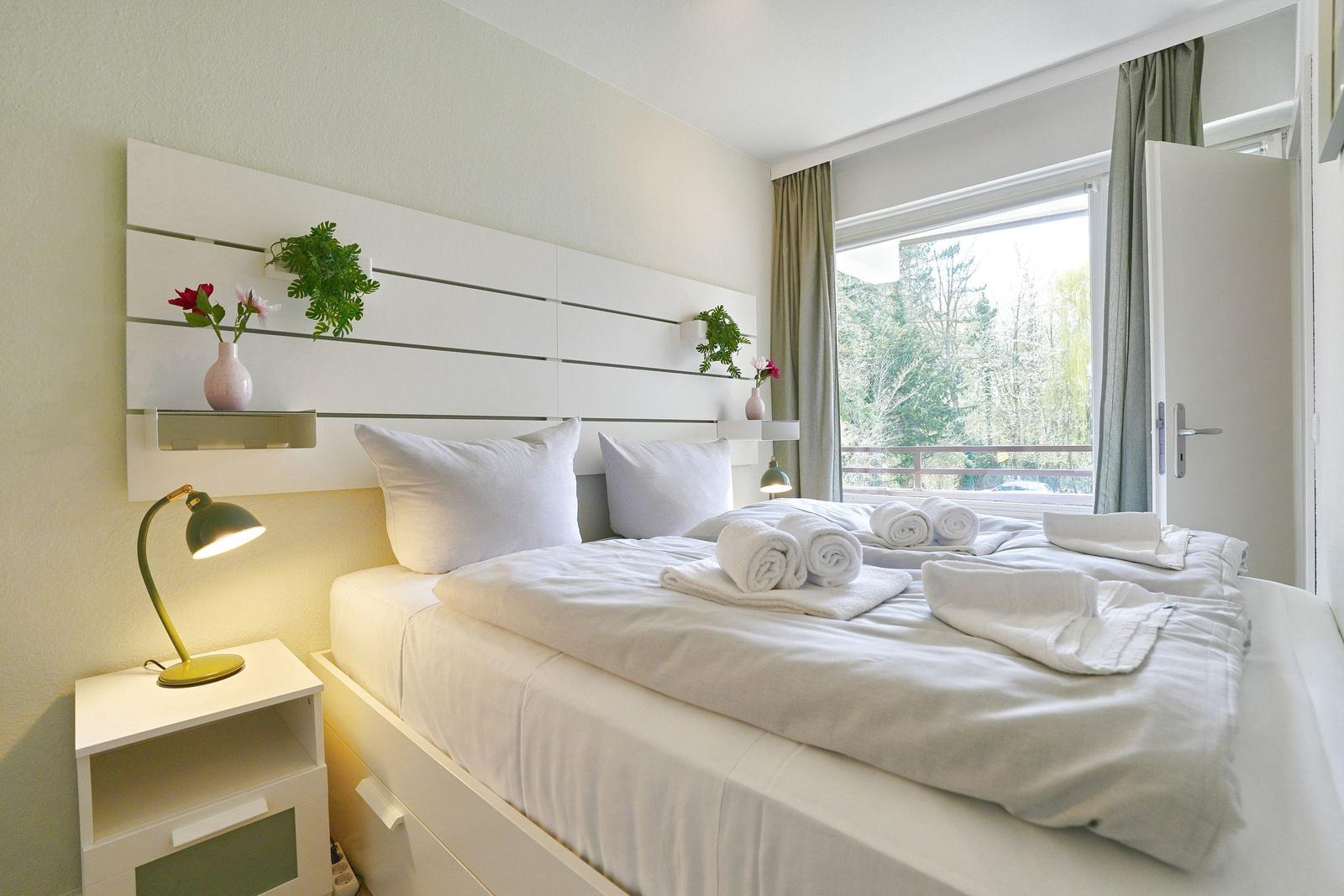 Schlafzimmer mit weißem Bett, Nachttisch und Fenster zum Balkon.