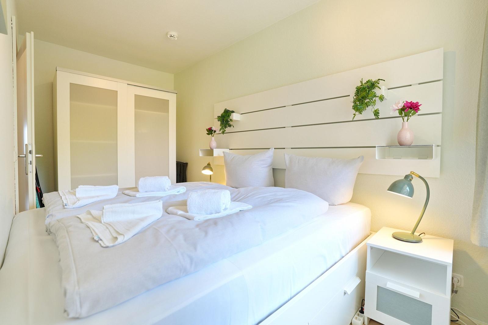 Ein Schlafzimmer mit weißem Bett, Nachttisch und Kleiderschrank. Auf dem Bett liegen Handtücher.