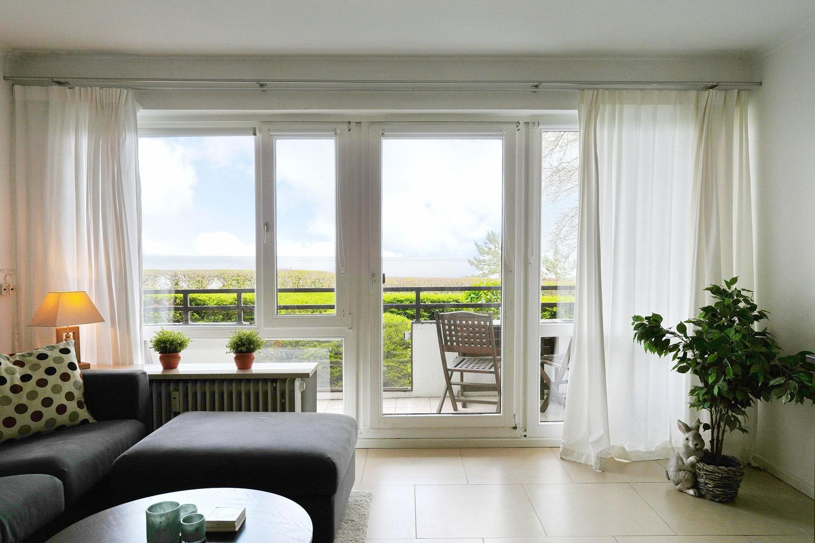 Wohnzimmer mit Blick auf Balkon und Grünfläche. Vorhänge, Sofa, Pflanzen und Tisch sind sichtbar.
