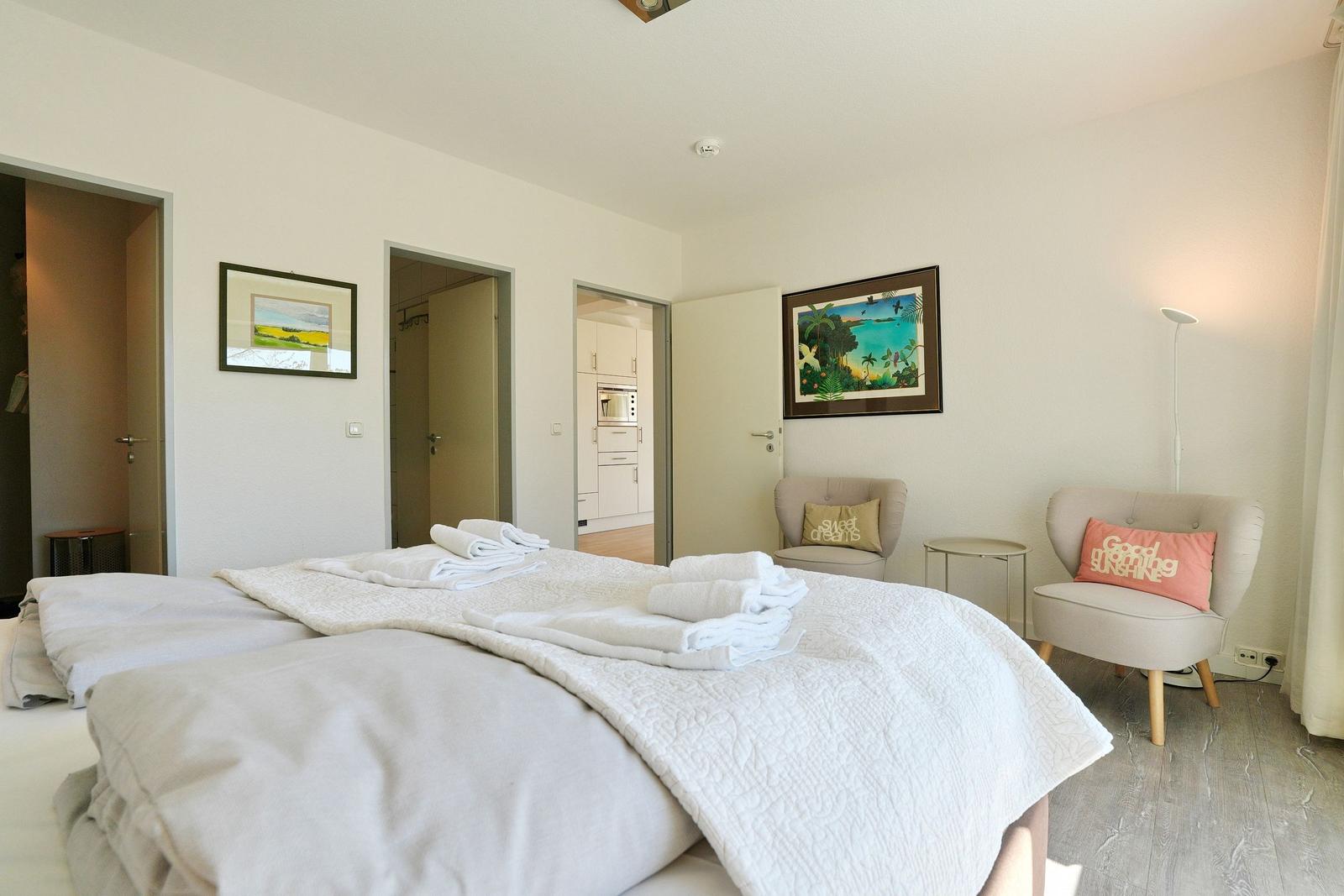 Schlafzimmer mit Bett, zwei Sesseln und Blick in die Küche.