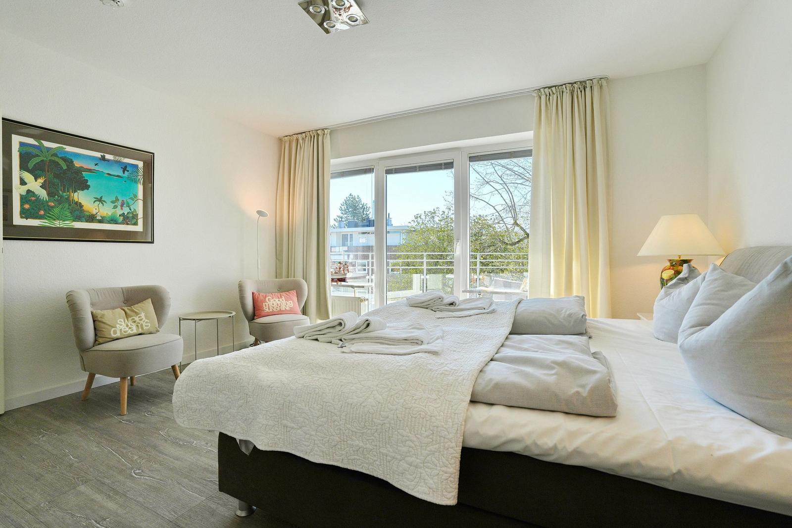 Schlafzimmer mit Bett, zwei Sesseln und Blick auf Balkon.