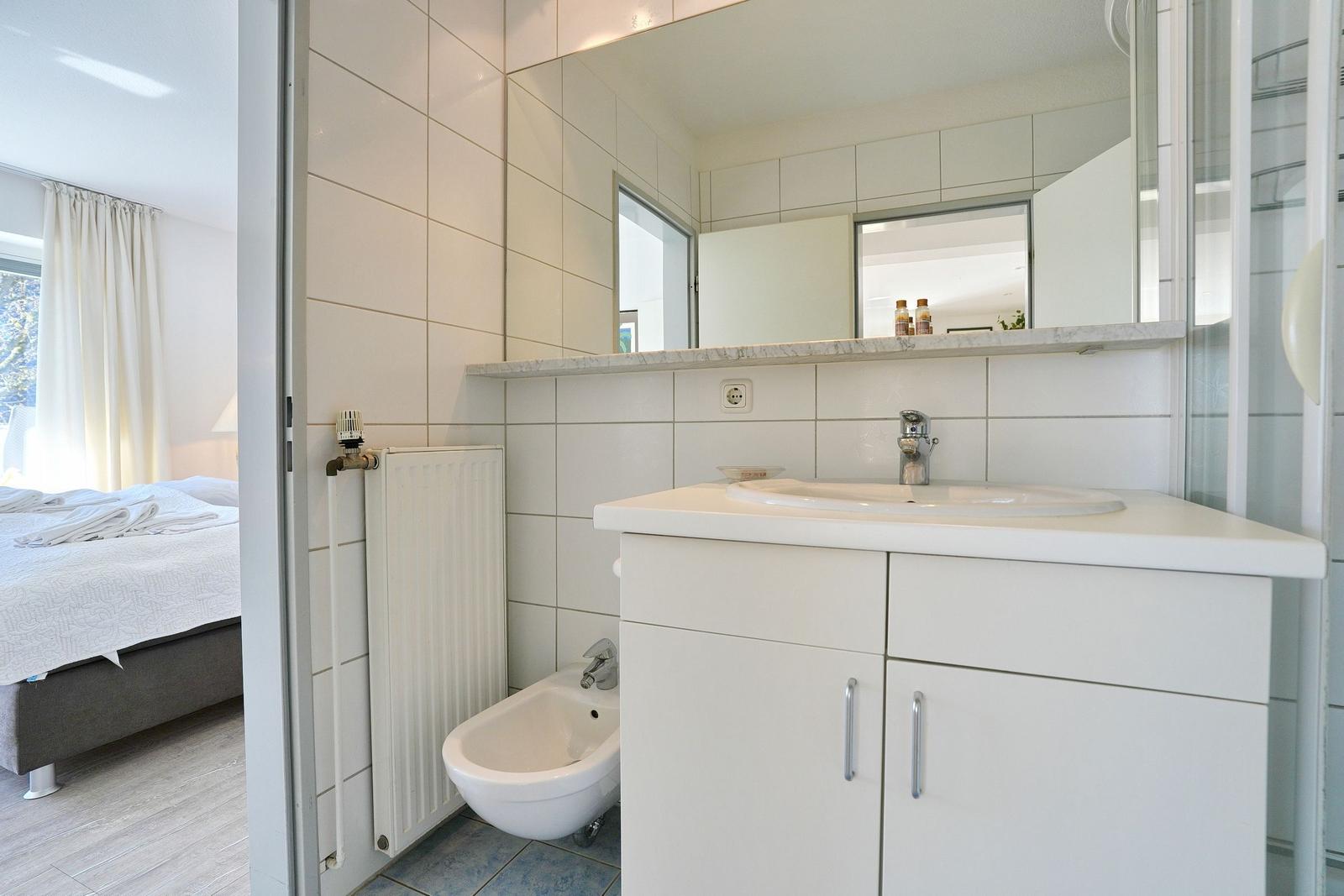 Badezimmer mit Waschbecken, Spiegel und Bidet. Blick in das Schlafzimmer mit Bett.