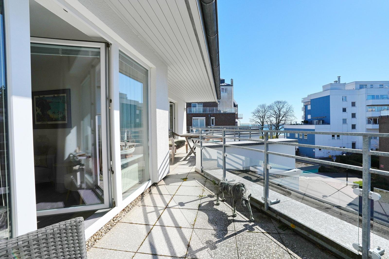 Terrasse mit Glasgeländer und Blick auf Gebäude. Im Hintergrund steht eine Statue.