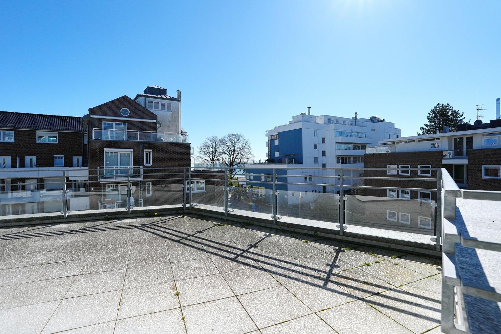 Terrasse mit Glasgeländer und Blick auf moderne Gebäude unter blauem Himmel.