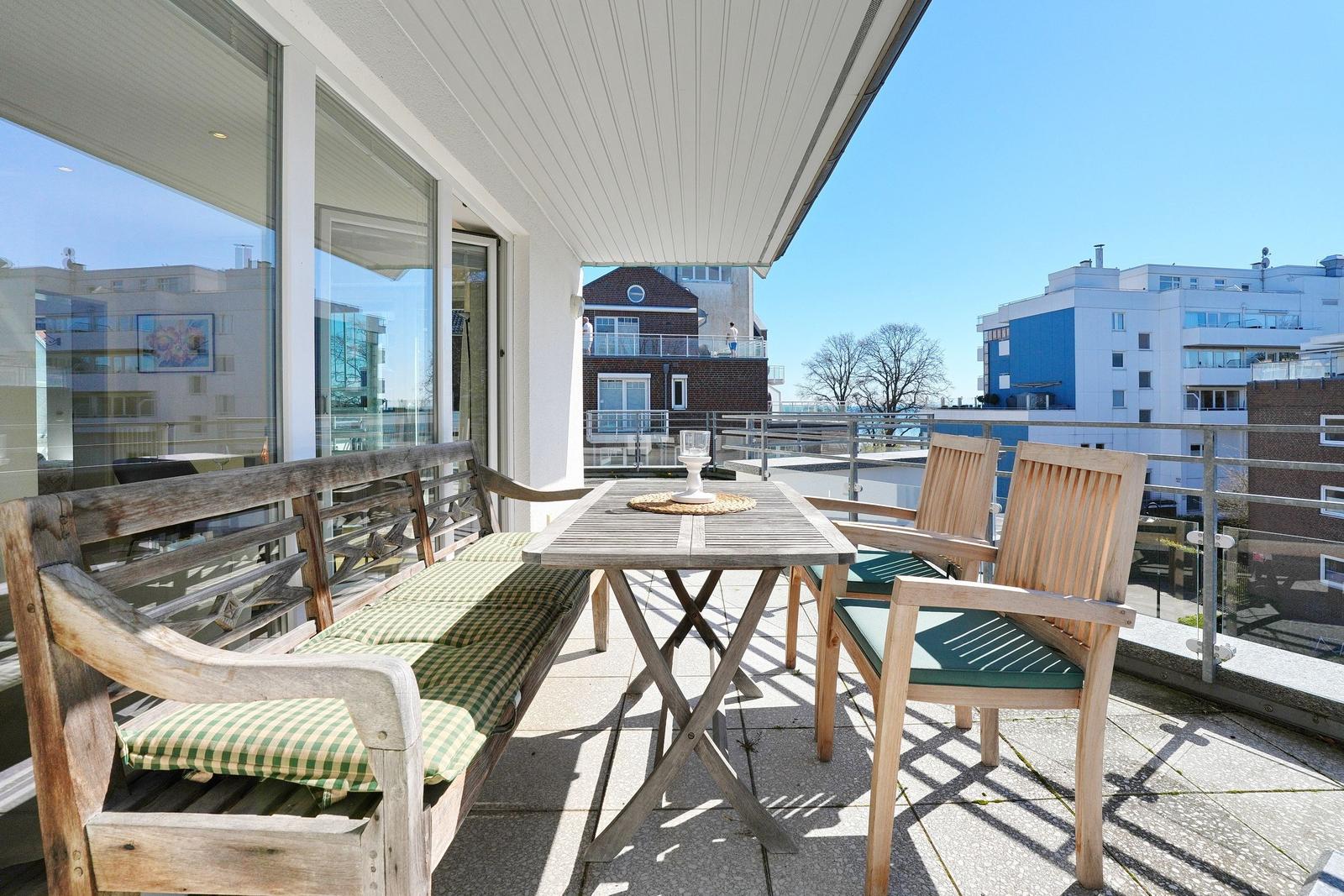 Terrasse mit Holztisch, zwei Stühlen und Bank mit Kissen. Blick auf Gebäude und klarer Himmel.