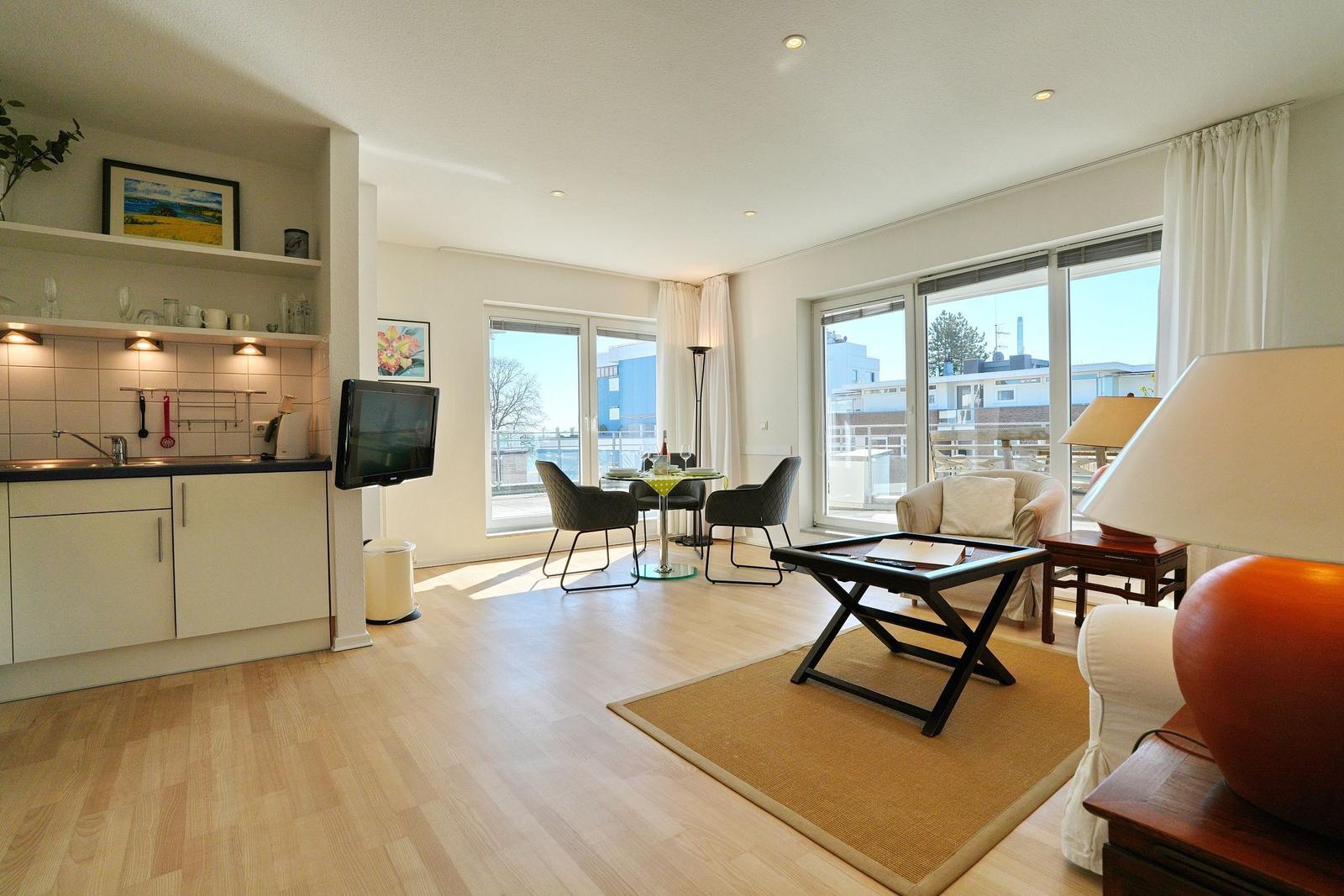 Wohnung mit Küche, Essbereich und Sitzgelegenheit. Große Fenster mit Blick auf Außenbereich.