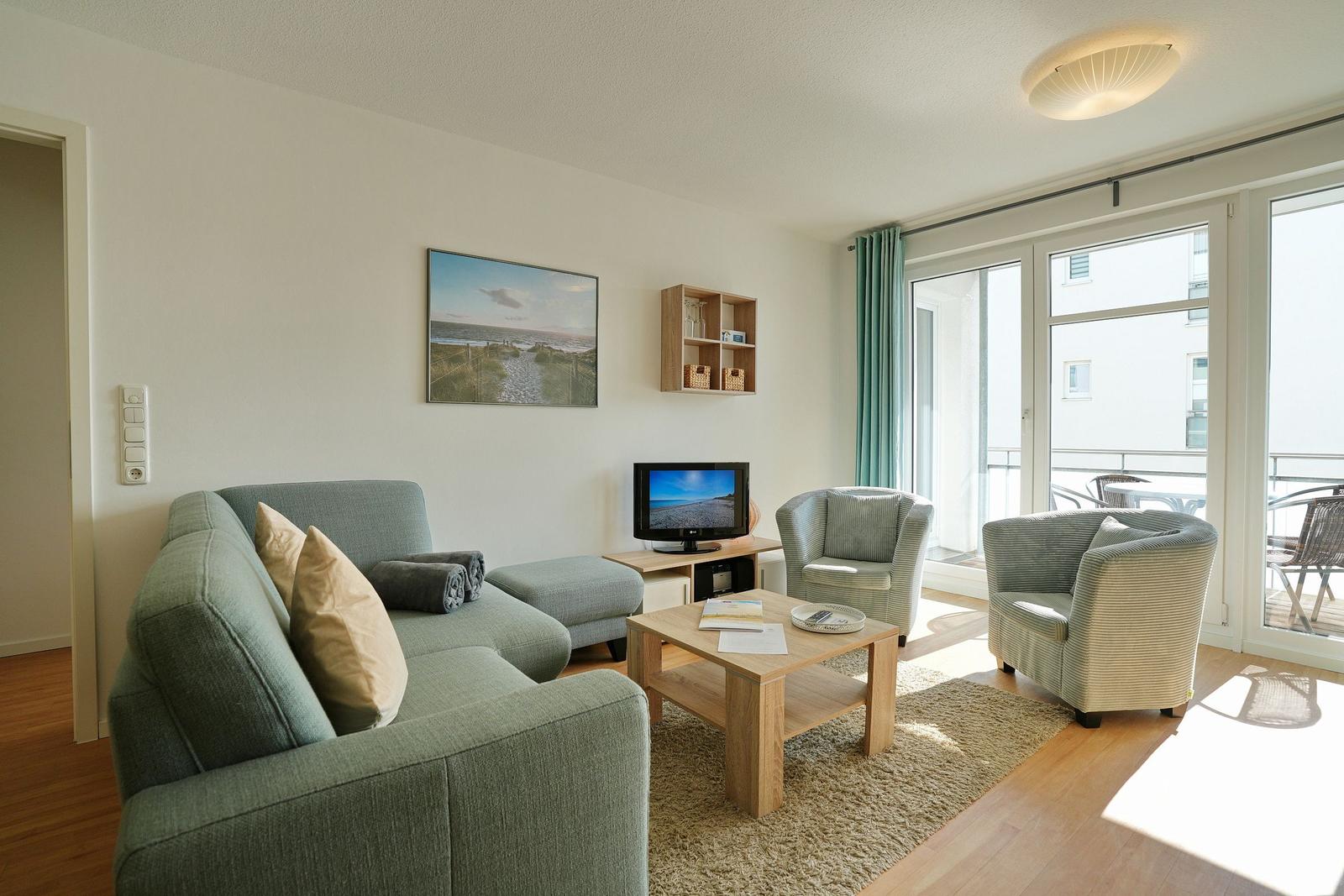 Wohnzimmer mit Sofa, zwei Sesseln, Fernseher und Balkonzugang.
