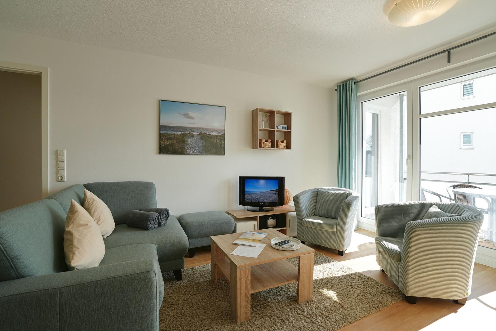 Wohnzimmer mit Sofa, zwei Sesseln, Couchtisch und TV. Große Fenster mit Blick auf Balkon.