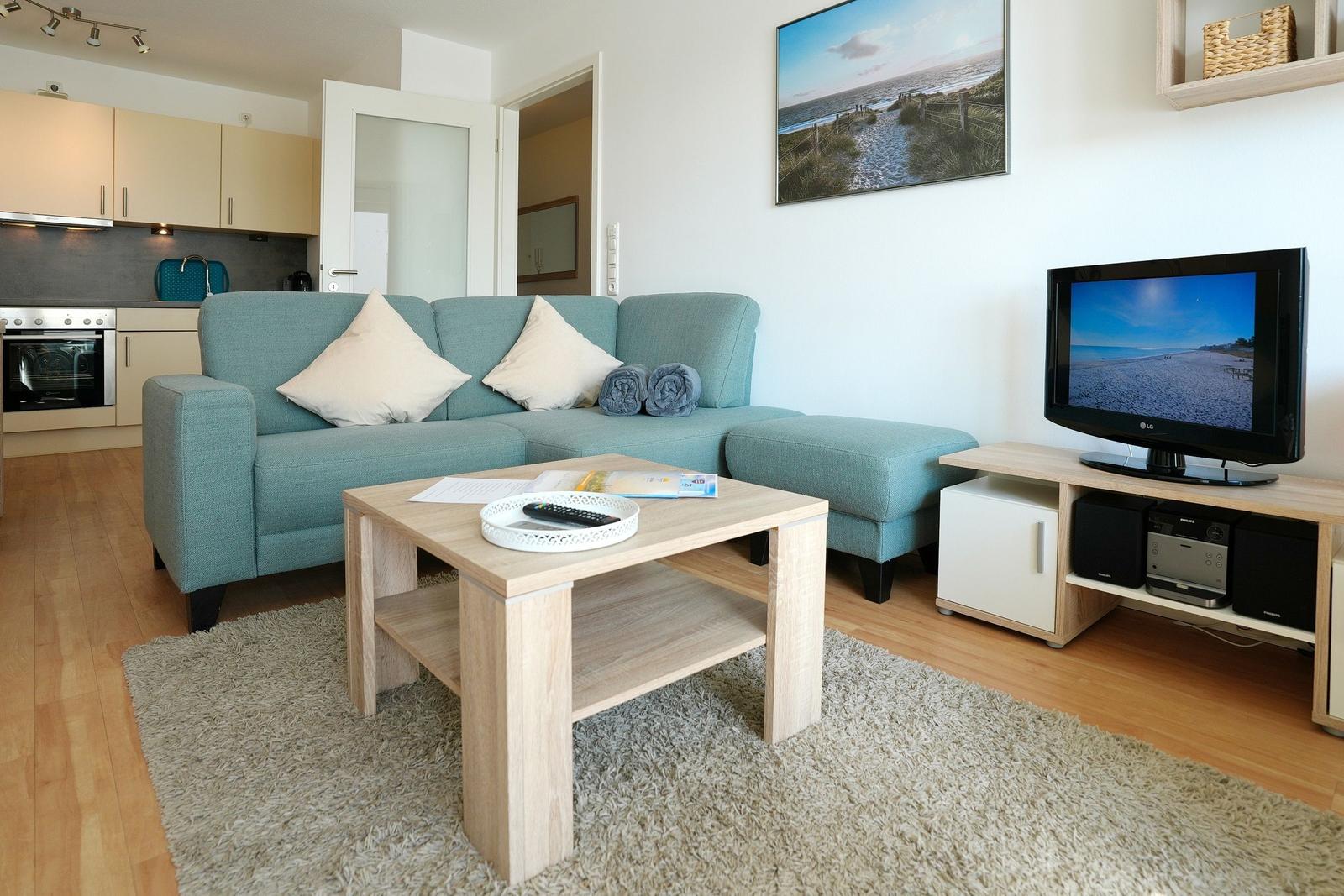 Wohnzimmer mit Sofa, Couchtisch und Fernseher. Küche im Hintergrund.