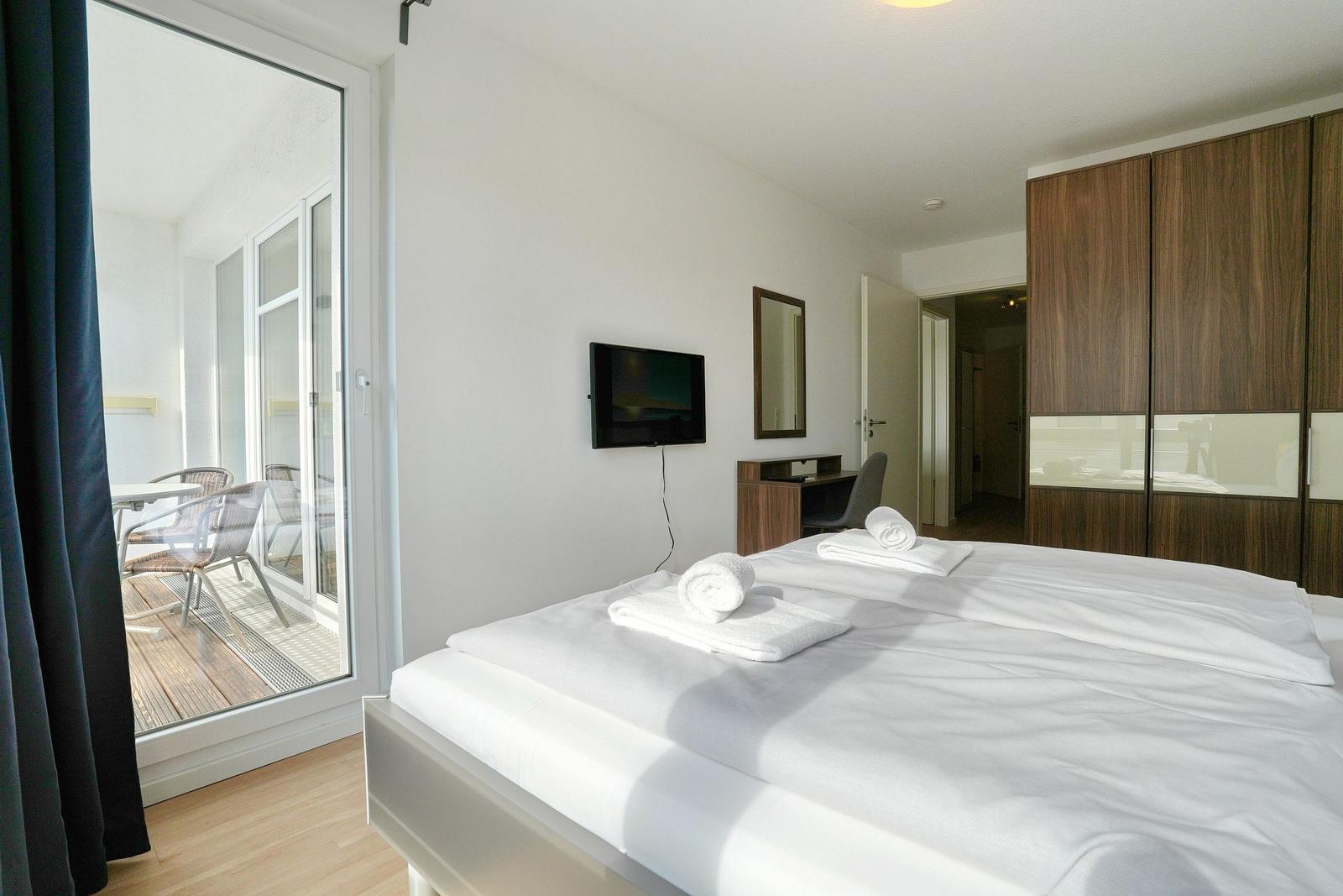 Schlafzimmer mit Doppelbett, TV und Balkon. Holzmöbel und weißer Bettwäsche.