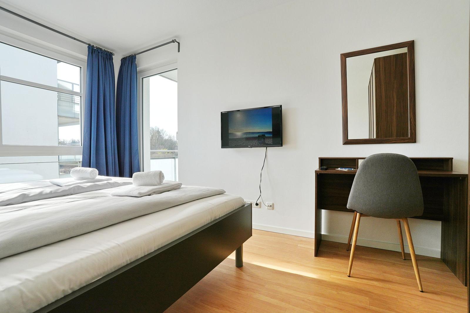 Schlafzimmer mit Doppelbett, Schreibtisch, Fernseher und Balkonansicht.