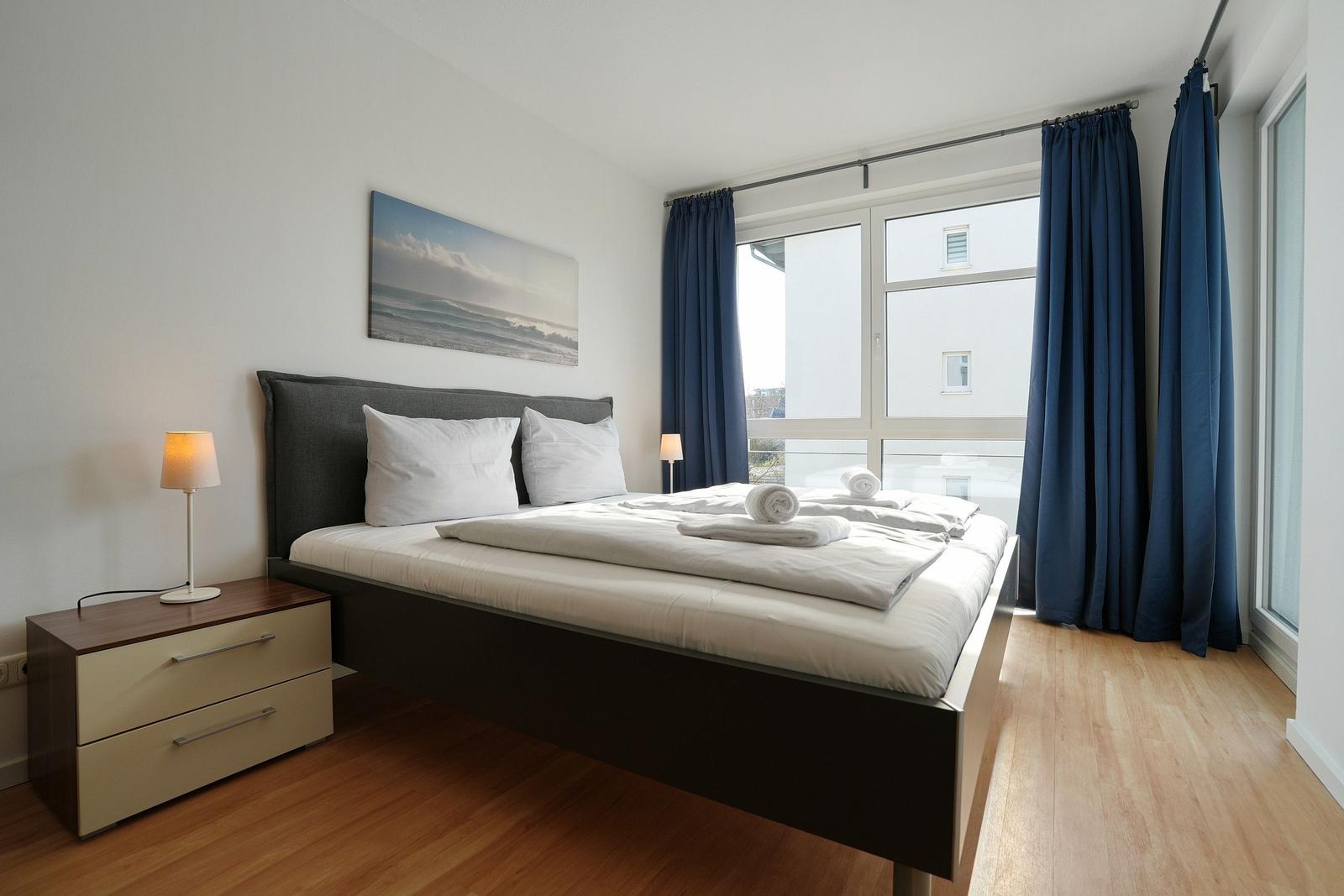 Schlafzimmer mit Bett, Nachttisch und Fenster mit blauen Vorhängen.