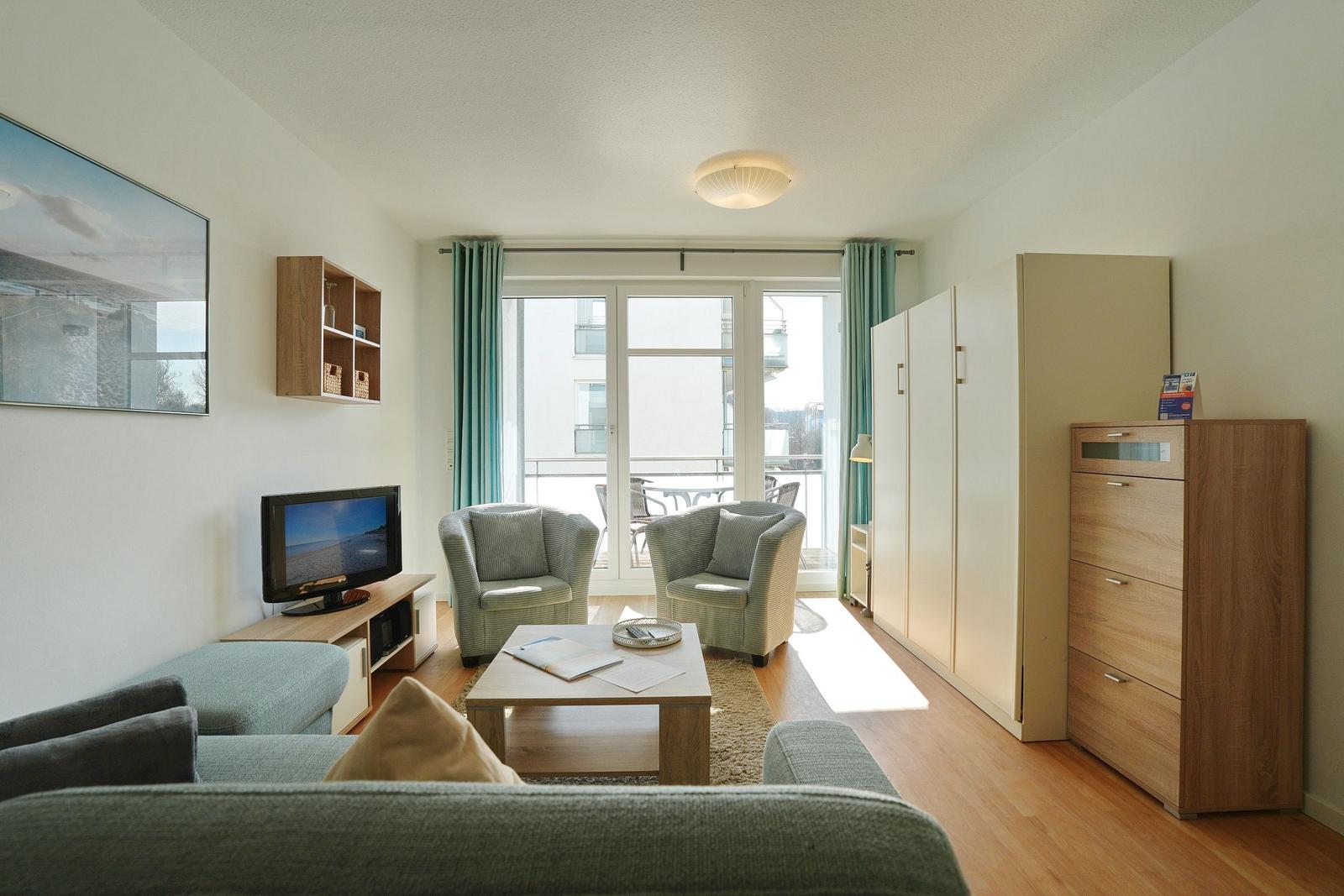 Wohnzimmer mit Sofa, zwei Sesseln, Fernseher und Balkonansicht.