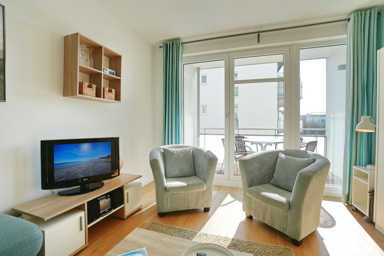 Wohnzimmer mit zwei Sesseln, Fernseher und Balkon. Holzboden und helle Wände.