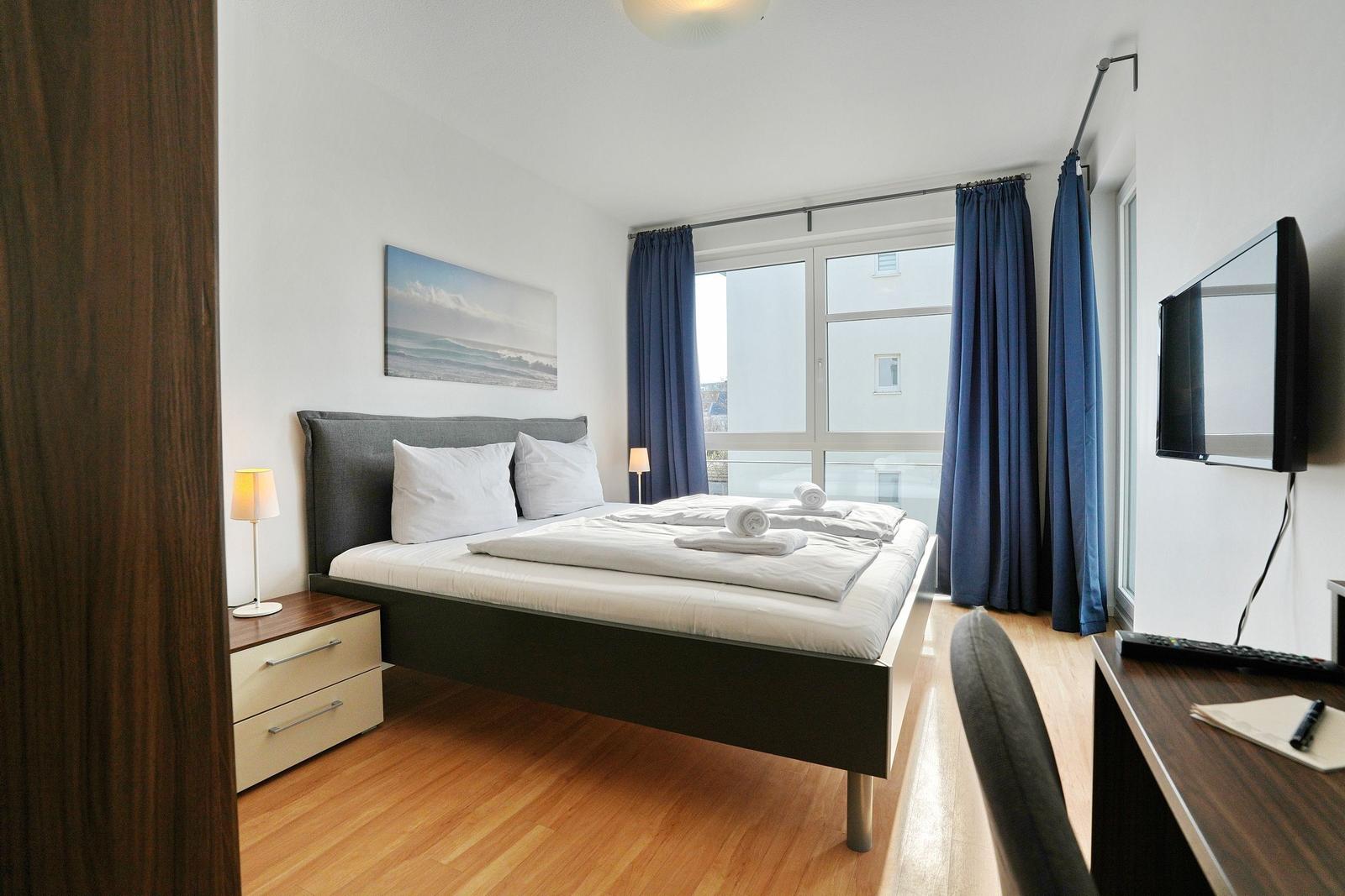Schlafzimmer mit Bett, Schreibtisch, Fernseher und Fenster mit blauen Vorhängen.