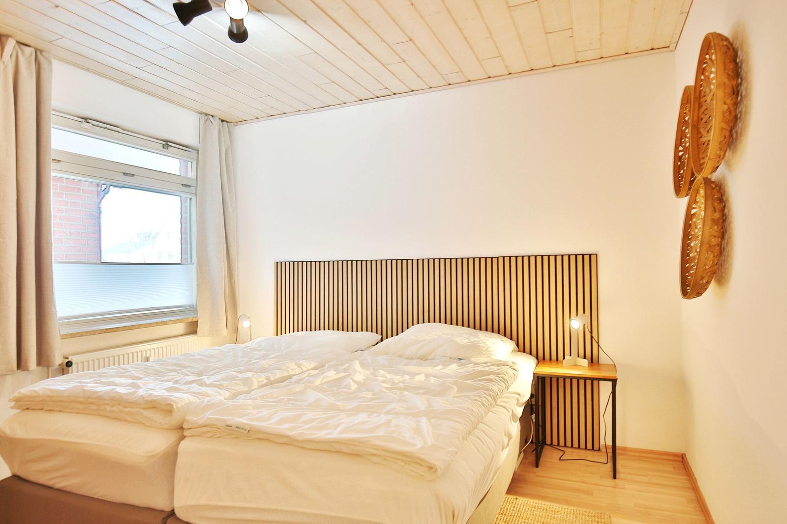 Doppelbett mit Holz-Kopfteil, Nachttisch und Fenster mit Vorhängen.