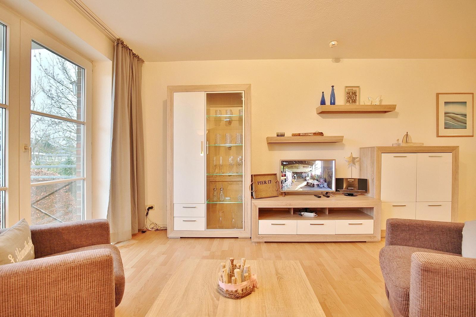 Wohnzimmer mit Holzmöbeln, Fernseher und Glasvitrine. Fenster mit Vorhang.