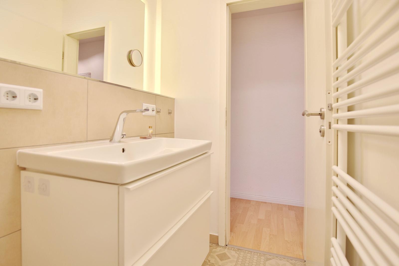 Badezimmer mit Waschbecken, Spiegel und Heizkörper. Tür führt in einen Raum mit Holzboden.