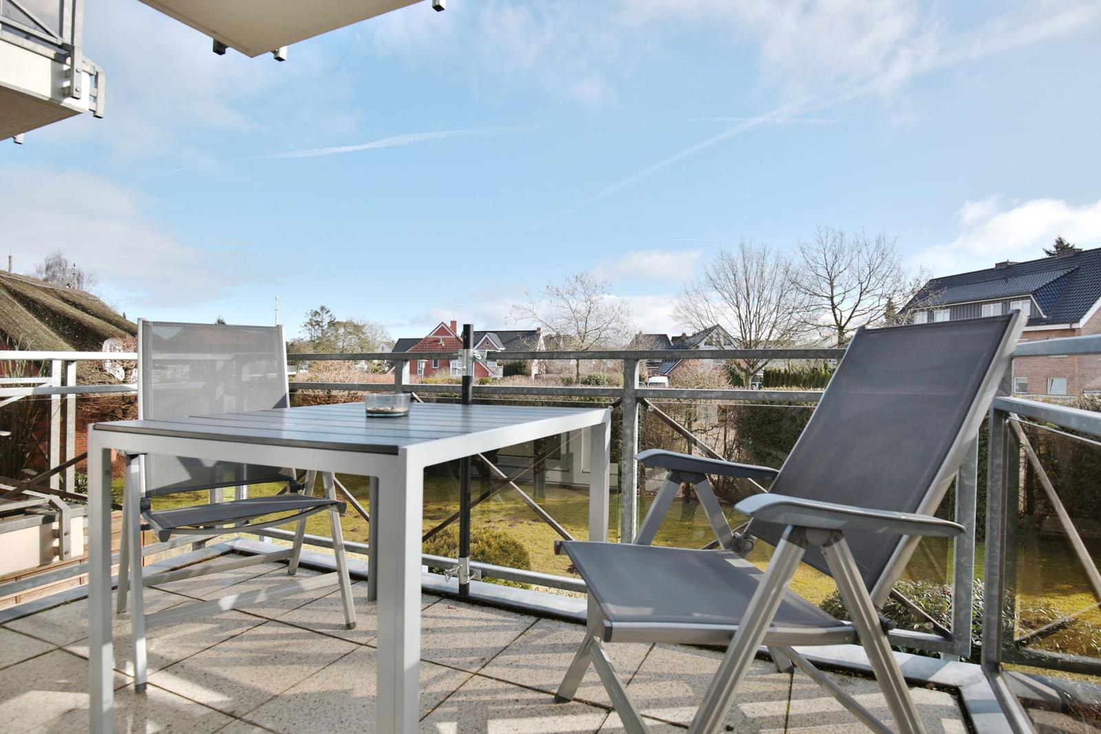 Terrasse mit Tisch und zwei Stühlen, Blick auf Wohnhäuser und Bäume unter blauem Himmel.